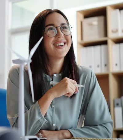 A young woman sits in front of a desk, holding a pen on her right hand and smiling. Next to her, there is a miniature version of a wind fan.
